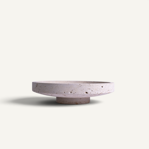A minimalist, low-profile round travertine bowl in a light grayish-lavender hue with natural dark speckles, set against a solid off-white background.