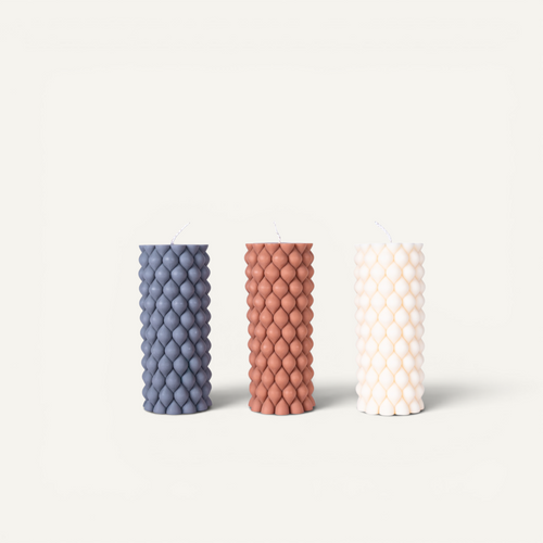 Three textured pillar candles standing upright in a row on a clean off-white background. The left candle is gray, the middle candle is terracotta, and the right candle is off-white.