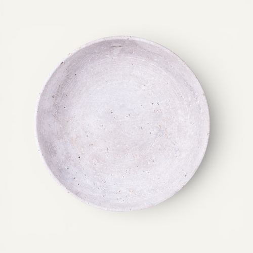 A round, shallow travertine bowl with a textured, speckled finish in a light lavender-gray color, photographed from directly above on a clean, off-white background.