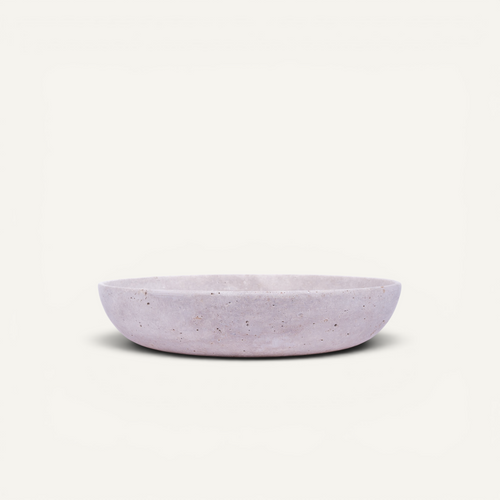 A shallow, oval-shaped travertine bowl in a light lavender purple color with subtle speckled texture, set against a solid off-white background.