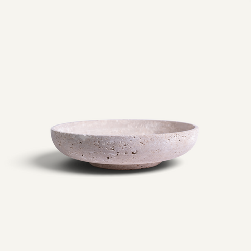 A shallow, wide travertine bowl with a textured, speckled finish in a muted dusty pink color, set against a plain off-white background.