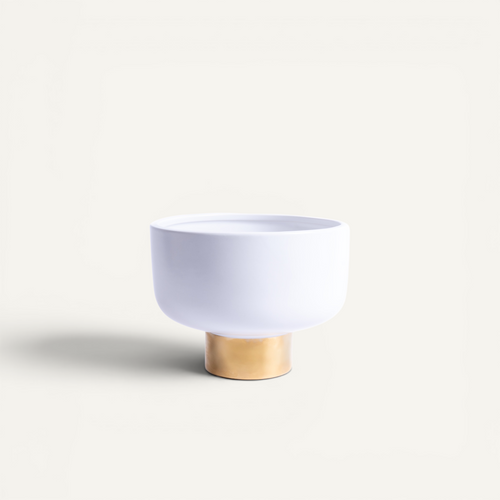 A minimalist white ceramic bowl with a flared rim, supported by a small cylindrical gold-toned base, set against a clean off-white background.