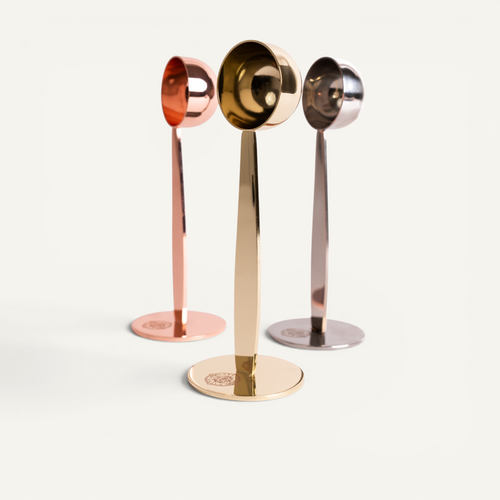 Three stainless steel espresso scoops in silver, copper, and gold colors with integrated tampers, displayed on a white background.