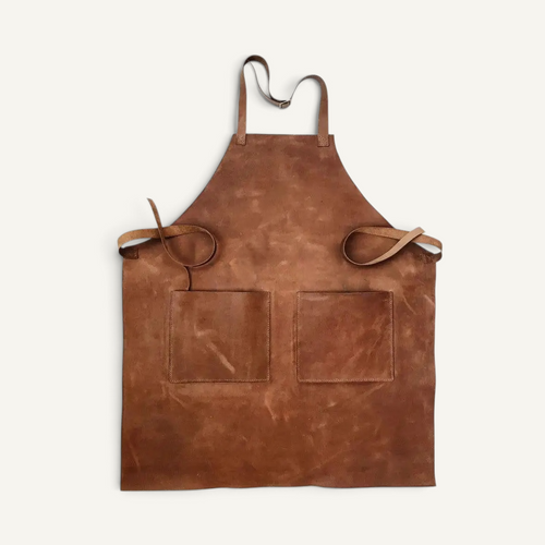A brown leather apron with two front pockets and adjustable neck and waist straps, laid flat against a light cream background.