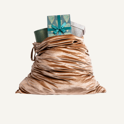 A large silk velvet Christmas sack in caramel color filled with wrapped gifts, including a green patterned box with a teal bow and a silver box, isolated on a light cream background.