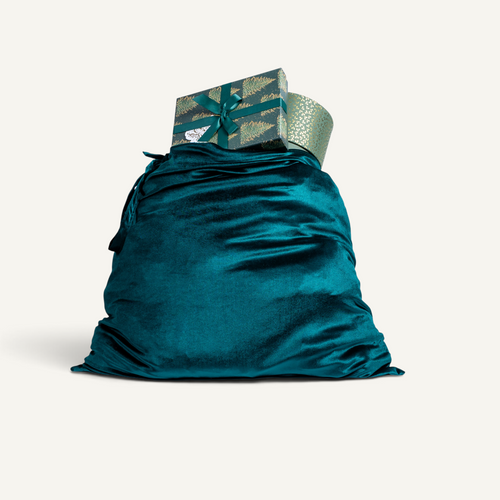 A large silk velvet Christmas sack in green color filled with wrapped gifts, including a green patterned box with a teal bow and a silver box, isolated on a light cream background.