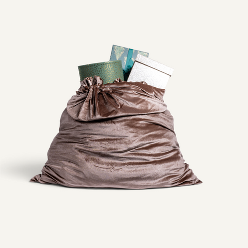A large silk velvet Christmas sack in caramel color filled with wrapped gifts, including a green patterned box with a teal bow and a silver box, isolated on a light cream background.