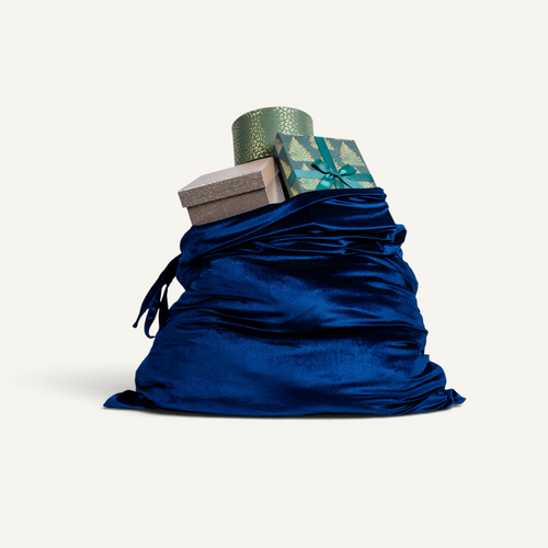 A large silk velvet Christmas sack in navy blue color filled with wrapped gifts, including a green patterned box with a teal bow and a silver box, isolated on a light cream background.