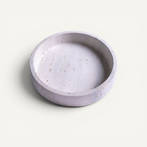 A round, shallow travertine bowl with a speckled light lavender-gray finish, photographed from a top-down angle on a clean, solid off-white background.