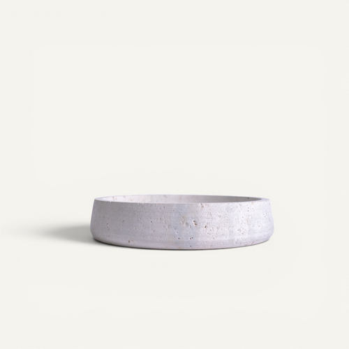 A minimalist, shallow round travertine bowl with a textured, speckled light gray finish, shown on a clean off-white background.
