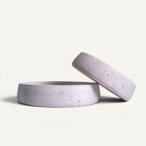 Two round, shallow travertine bowls both in a light lavender-gray color with subtle speckled texture, set against a solid off-white background.