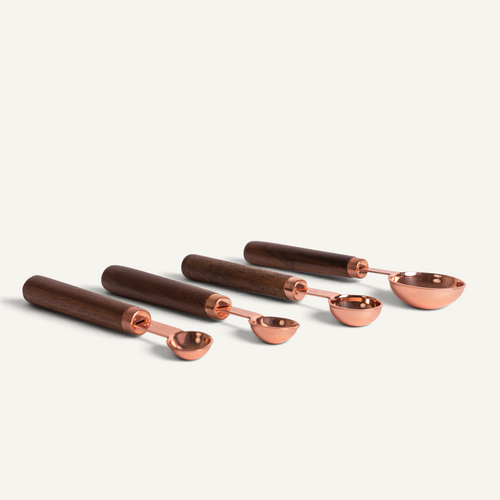 Four measuring spoons with walnut handles and polished copper-colored metal bowls, arranged diagonally on a light off-white background.