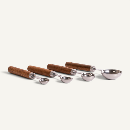 Four metal measuring spoons with brown wooden handles, arranged in a row on a light cream background.