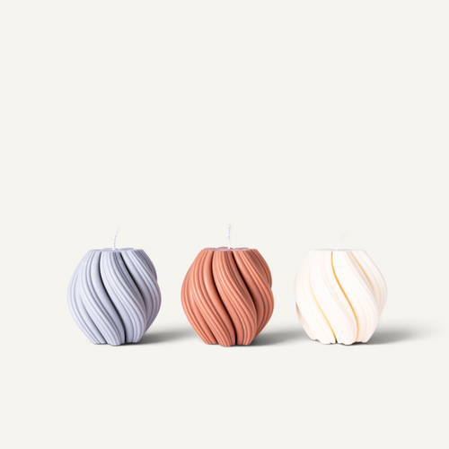 Three ribbed, rounded candles in pastel shades of lavender, terracotta, and cream, arranged side-by-side on a light beige background.