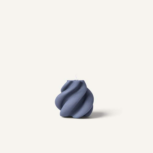 A modern, sculptural ribbed candle holder in a muted slate blue-gray color, featuring a wavy, organic shape and a single white wick at the top, set against a solid off-white background.