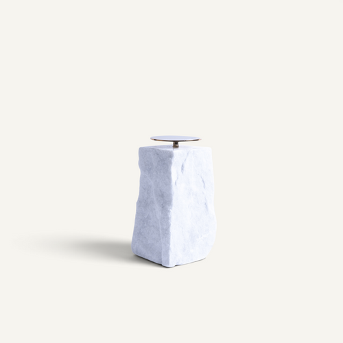 A modern minimalist candle holder featuring a textured, light blue-gray stone pedestal base with a small, circular metallic dish on top, set against a solid off-white background.